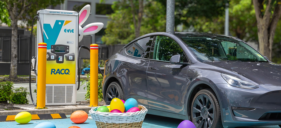 An Evie EV charger with white bunny ears next to a grey electric car, with colourful Easter eggs and a basket scattered in the parking bay